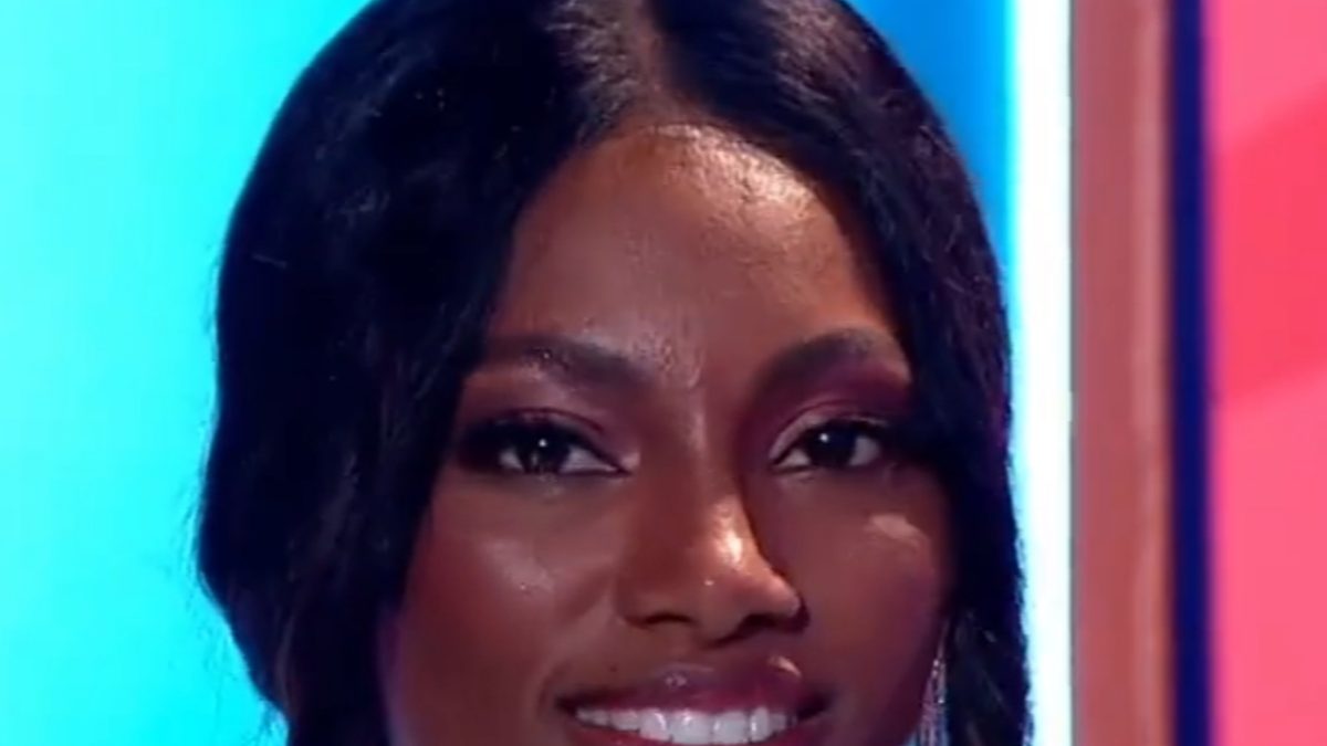 Tina Calamba sorrindo em close, em estúdio de TV com fundo azul e vermelho, em imagem de 2023