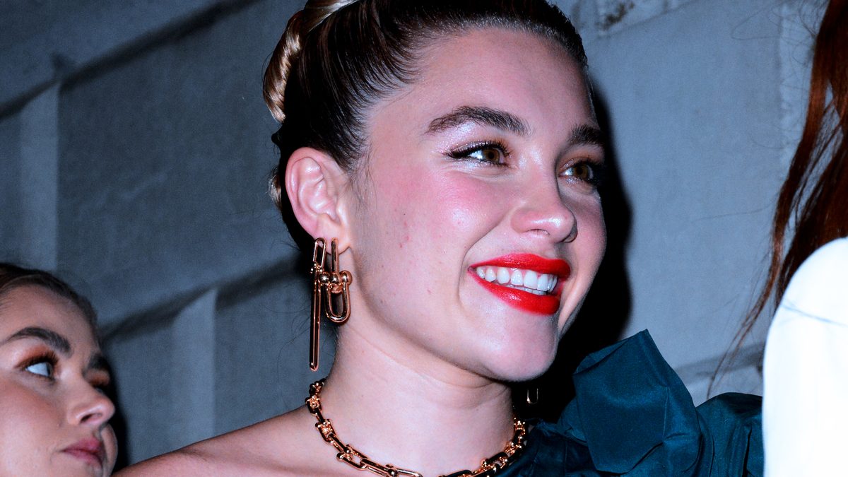 A atriz Florence Pugh sorri, de vestido azul de um ombro só e batom vermelho, em uma festa em 2020