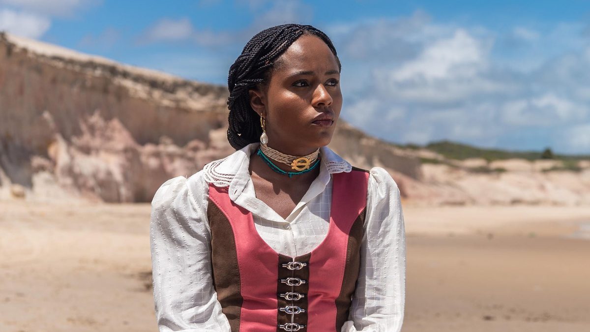 A atriz Duda Santos, como a princesa Alika em A Nobreza do Amor, sentada numa praia de falésias, olha séria para o lado em foto de divulgação