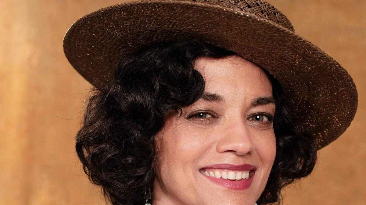 A atriz Ana Cecília Costa, como Teresa em A Nobreza do Amor, de chapéu de palha e vestido estampado, sorri para a câmera em foto de divulgação