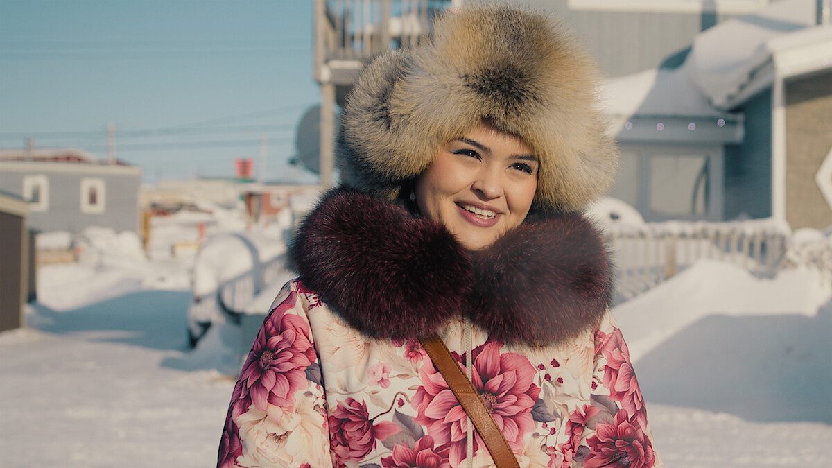 Anna Lambe, intérprete de Siaja, sorri com gorro e gola grandes de pele e casaco florido, numa rua coberta de neve, em imagem de divulgação da 2ª temporada de Ao Norte do Norte