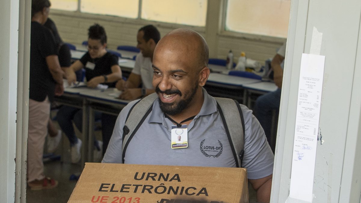 Funcionário de camisa cinza e crachá sorri ao carregar a caixa de uma urna eletrônica na porta de uma seção eleitoral, no segundo turno das eleições de 30 de outubro de 2022