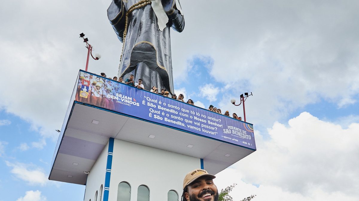 Paulo Vieira, de camisa estampada e mala de couro na mão, diante de um monumento branco com estátua