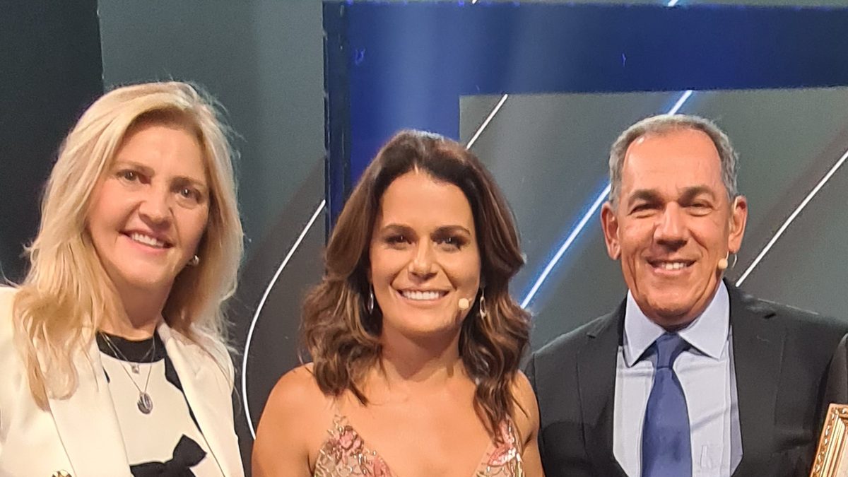 A jornalista Adriana Araújo, ao centro, sorri no palco do Prêmio Band Cidades Excelentes, em novembro de 2023, ao lado de dois participantes da cerimônia