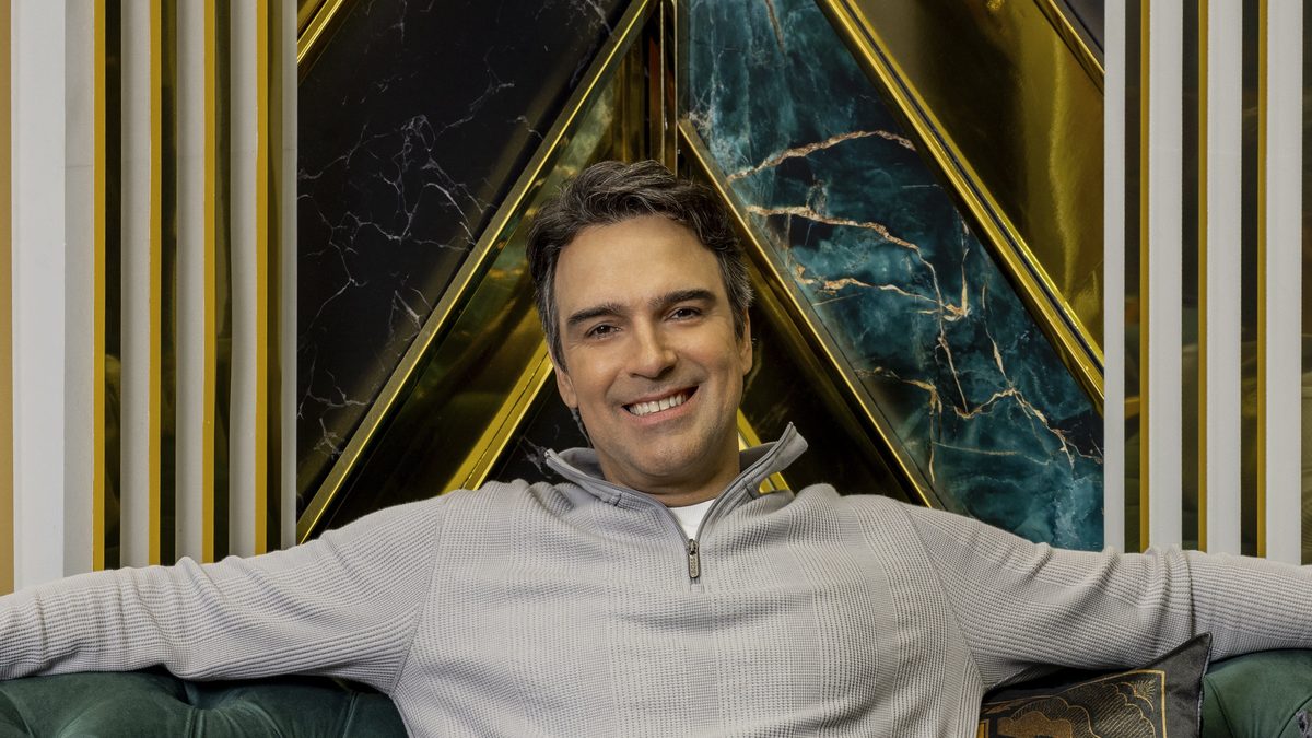 Tadeu Schmidt sorri de braços abertos, sentado num sofá verde na casa do BBB 26, em foto de divulgação da TV Globo feita em janeiro de 2026