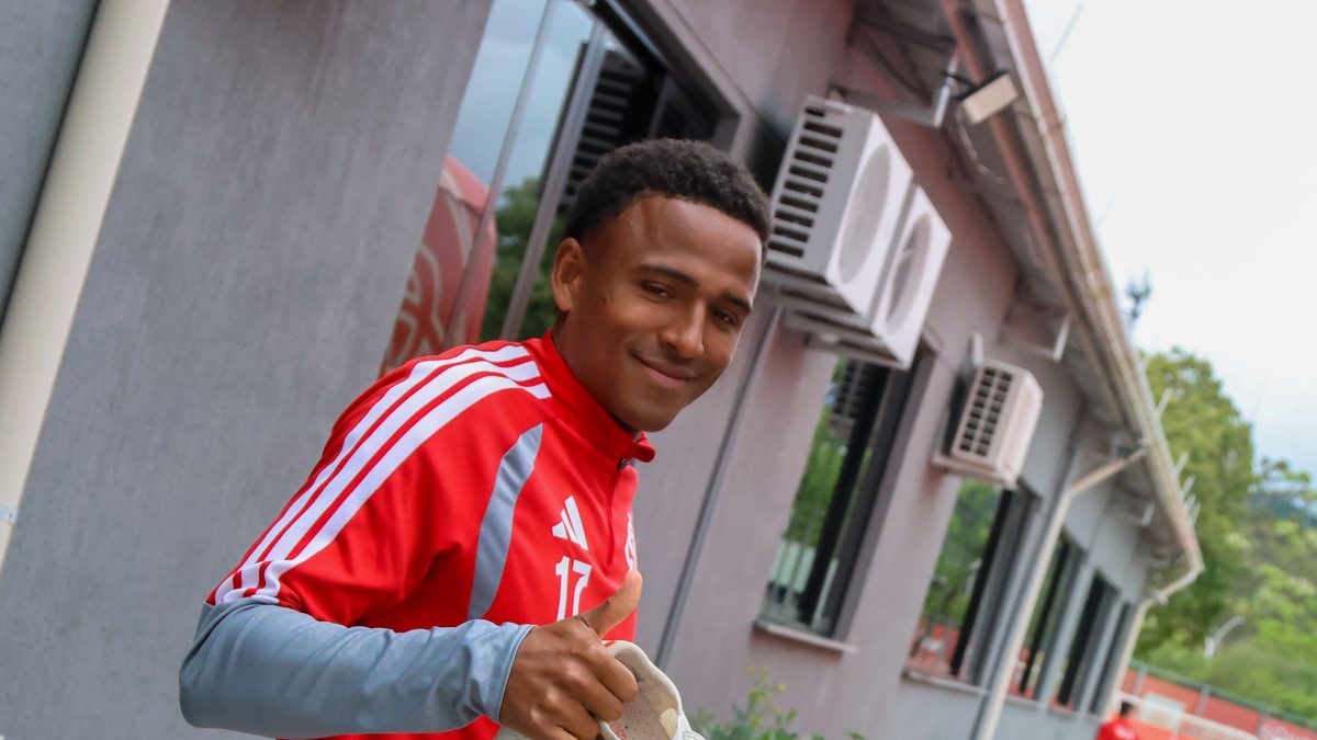 Jogador do Internacional, de uniforme de treino vermelho com o número 17, sorri e faz sinal de positivo com as chuteiras na mão, no centro de treinamento do clube, na preparação para enfrentar o Corinthians