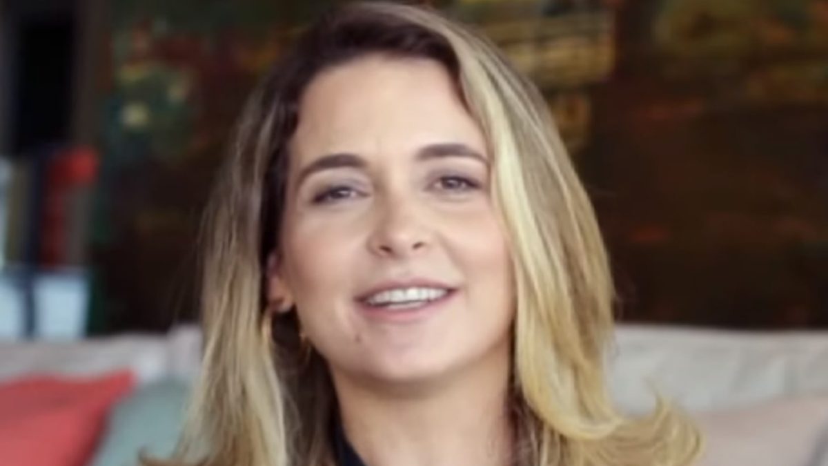 A atriz Cláudia Abreu sorri, de cabelo loiro e blusa escura estampada, sentada numa sala, em 2019