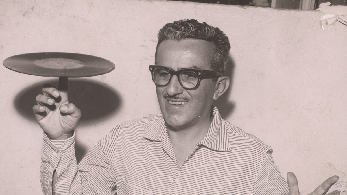 Chacrinha, de óculos de aro grosso e bigode, sorri e equilibra um disco de vinil na ponta do dedo, em foto de 1961