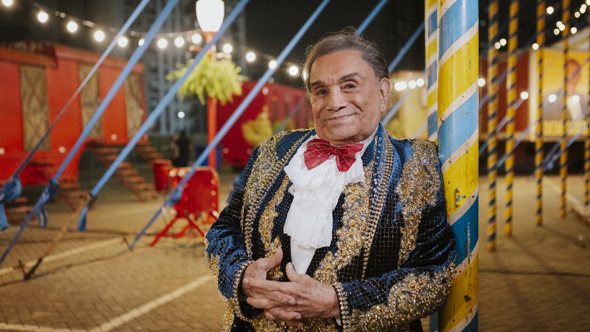 Dedé Santana, de casaca azul e dourada e gravata borboleta vermelha, sorri encostado em um mastro de circo, em turnê pelo Brasil