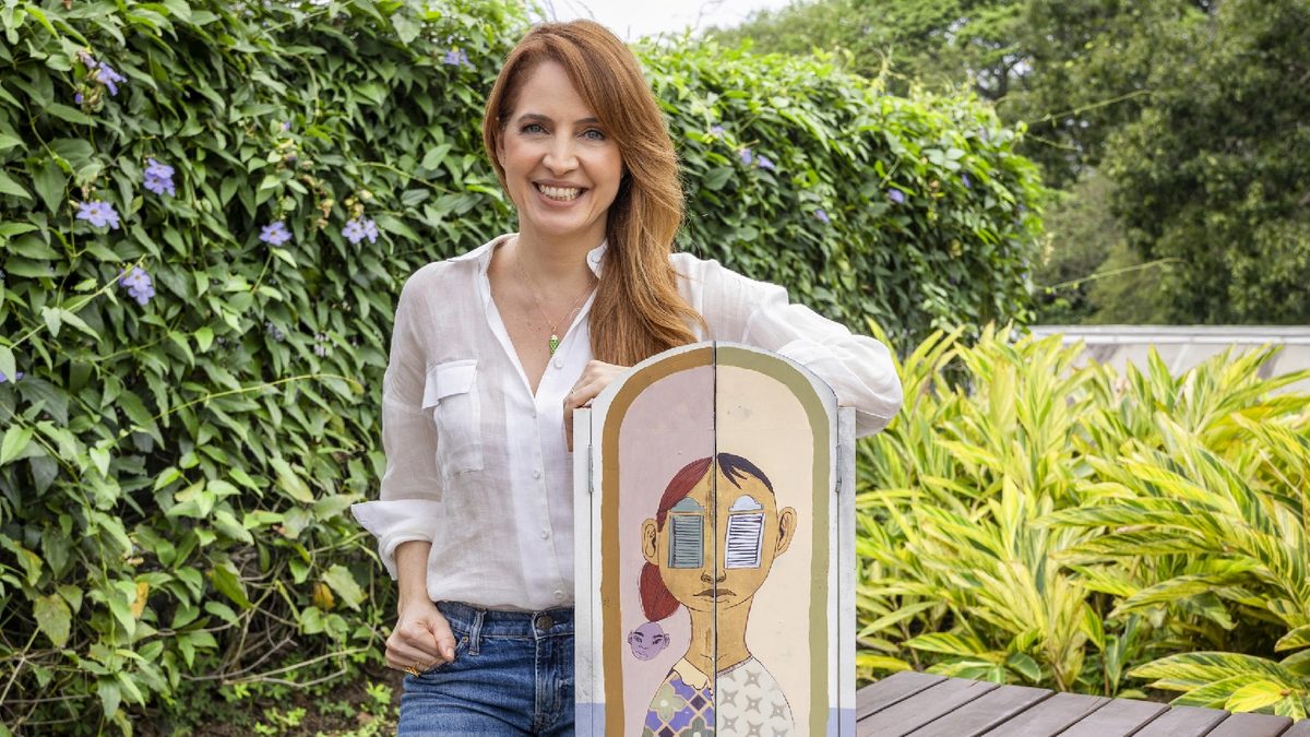 Poliana Abritta, de camisa branca e jeans, sorri e segura uma peça de arte com o desenho de um adolescente, num jardim