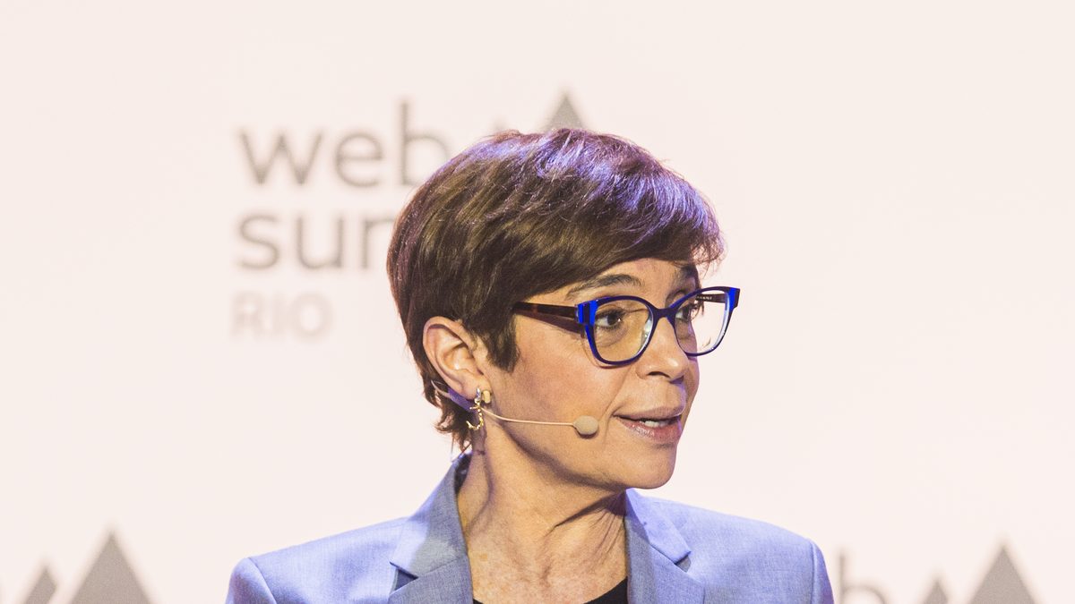 A jornalista Renata Lo Prete, de terno azul-claro e óculos, fala sentada em uma poltrona branca durante o Web Summit Rio, em 2024
