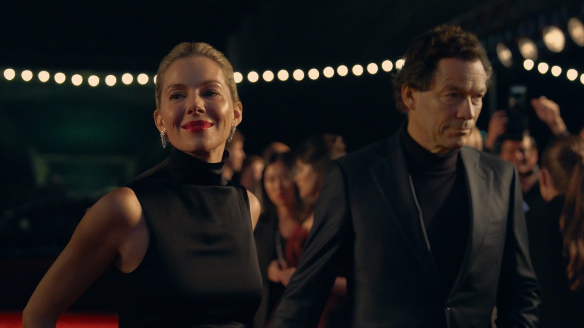 Sienna Miller, de vestido preto e batom vermelho, e Dominic West, de terno escuro, num tapete vermelho em cena de WAR