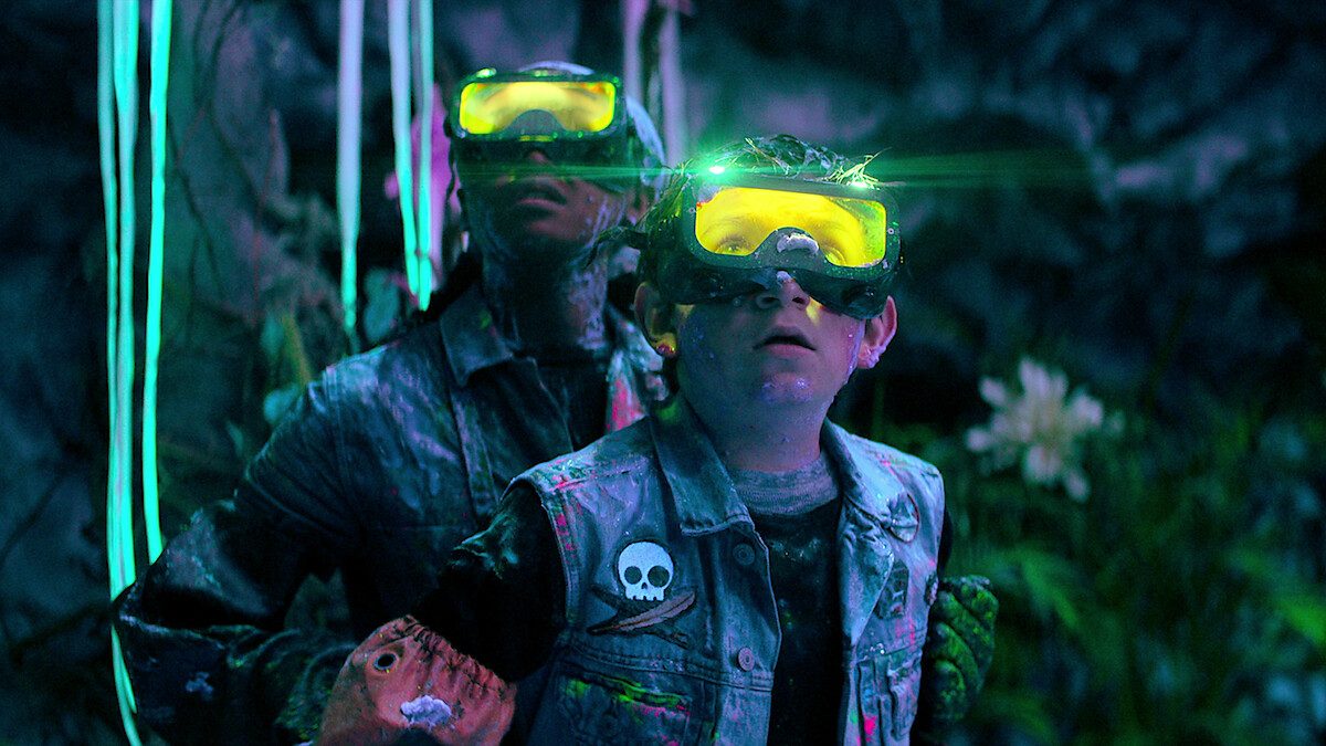Dois jovens de óculos de proteção brilhantes e jaquetas jeans numa caverna iluminada por neon, em imagem de divulgação da Netflix