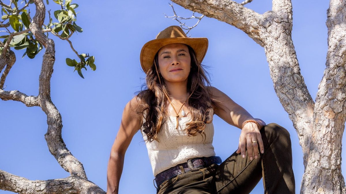 Dira Paes, de chapéu, sentada no galho de uma árvore com a Chapada Diamantina ao fundo, em foto de divulgação de Rosenda em Lá na Minha Terra