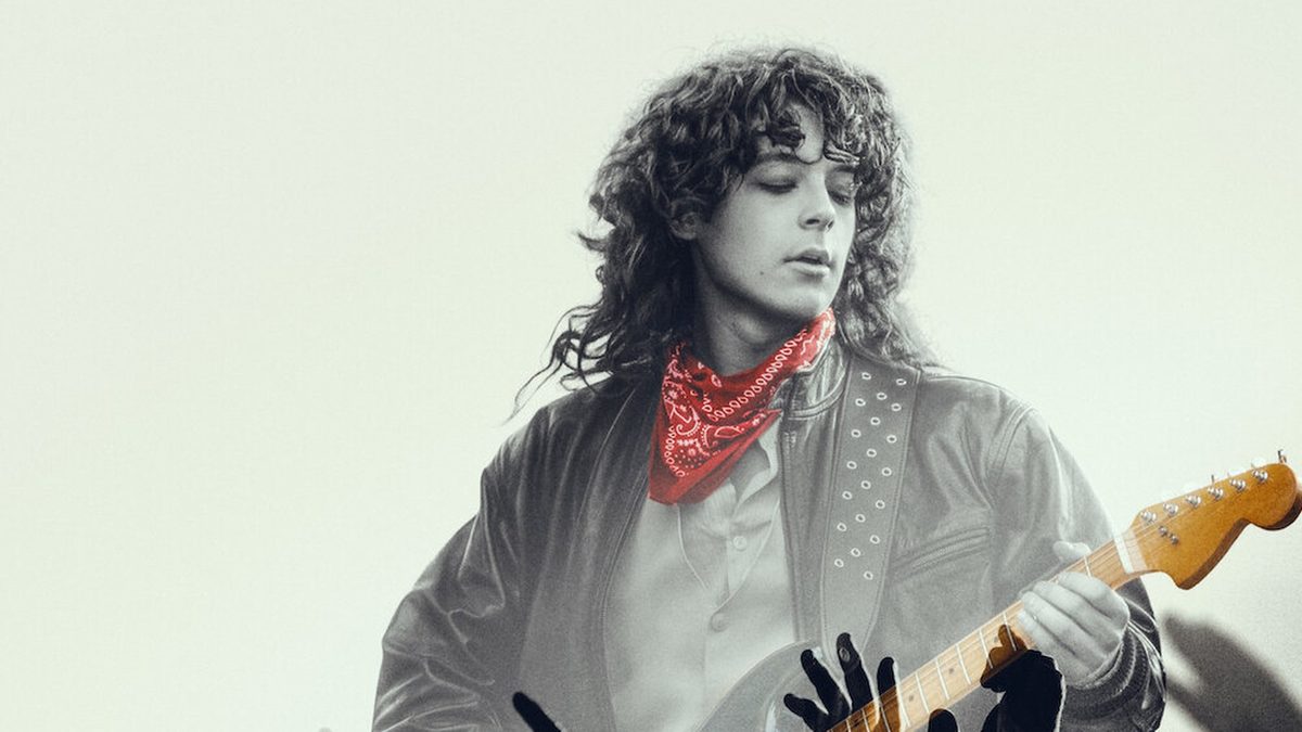 Jovem de cabelo cacheado e lenço vermelho no pescoço toca uma guitarra vermelha, em imagem de divulgação de Less of a Stranger na Netflix