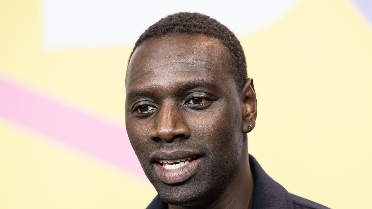 O ator Omar Sy, de jaqueta azul-marinho, sorri diante de um painel amarelo no Festival de Berlim de 2020