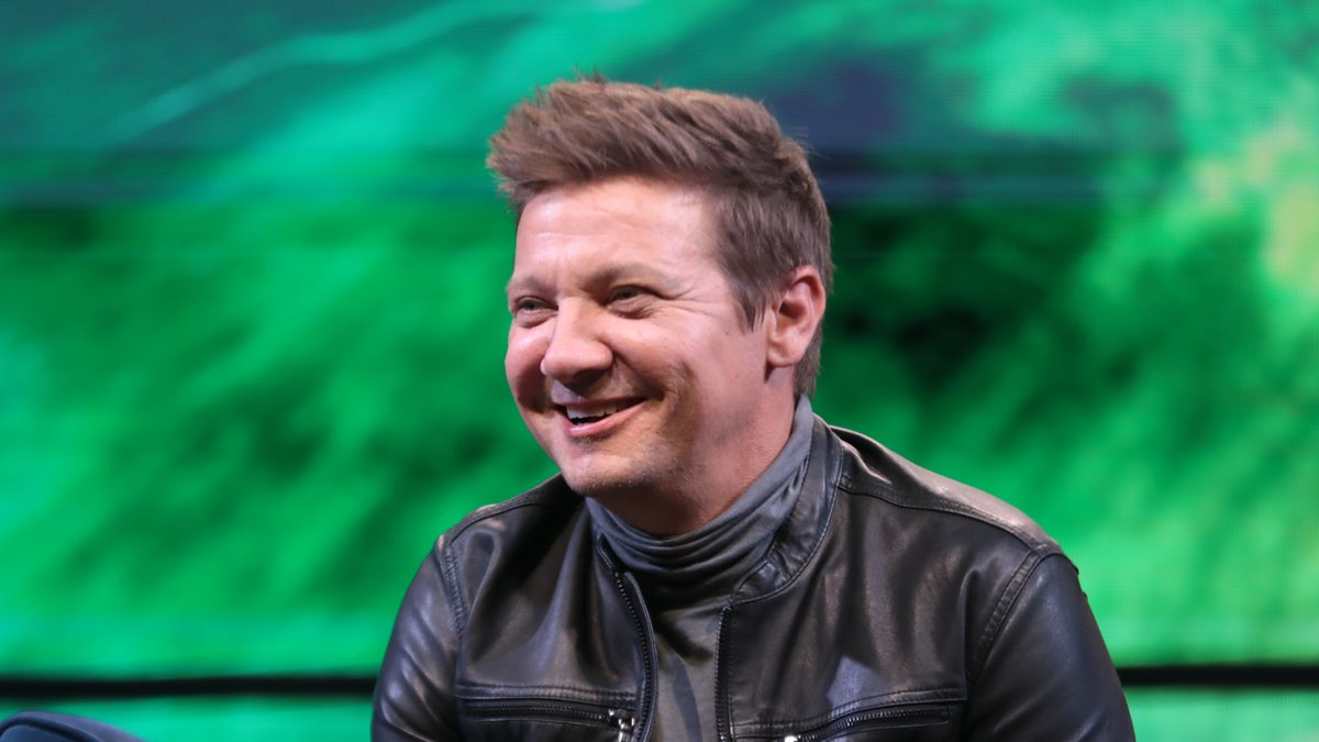 O ator Jeremy Renner, de jaqueta de couro preta, sorri com um microfone na mão durante painel na Phoenix Fan Fusion, em Phoenix (Arizona), em junho de 2023