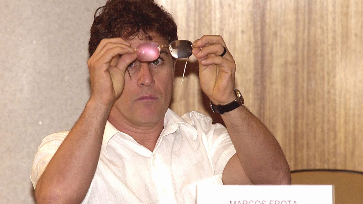 Marcos Frota segurando um par de óculos rosa diante do rosto, de camisa branca