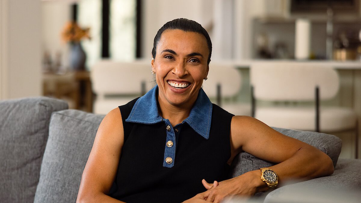 Marta sorri sentada em um sofá, de camisa preta com gola azul e relógio dourado, em foto de divulgação do documentário da Netflix