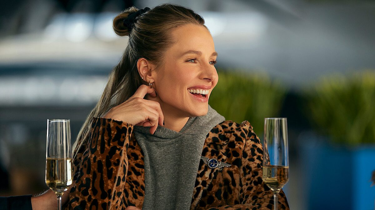 Kristen Bell, a Joanne de Ninguém Quer, sorrindo entre duas taças de champanhe em imagem de divulgação da 3ª temporada da série da Netflix