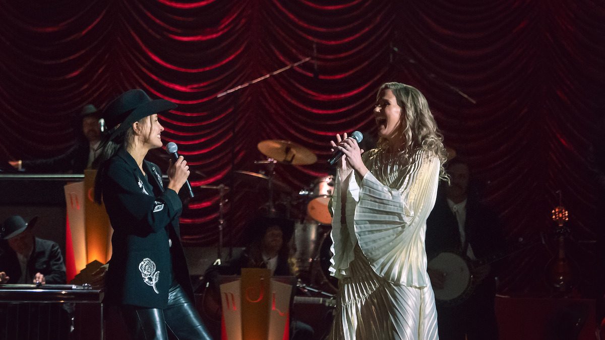 Cena de No Coração de Nashville, da Netflix: duas cantoras dividem o palco, uma delas de chapéu e jaqueta preta, diante de cortina vermelha