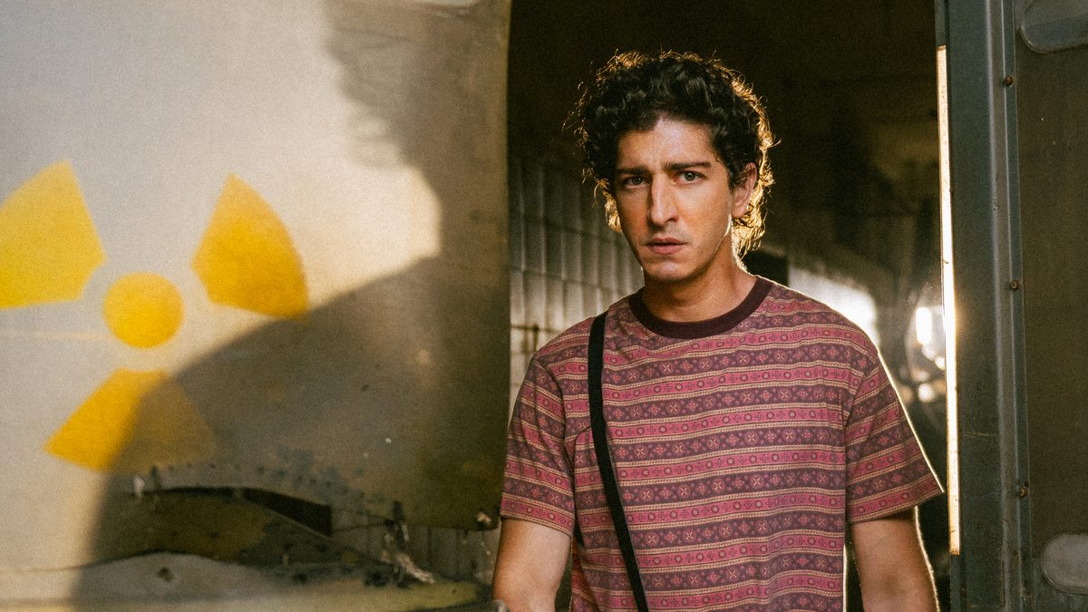 Johnny Massaro como Márcio em Emergência Radioativa, ao lado de uma parede com o símbolo amarelo de radiação, em foto de divulgação da Netflix