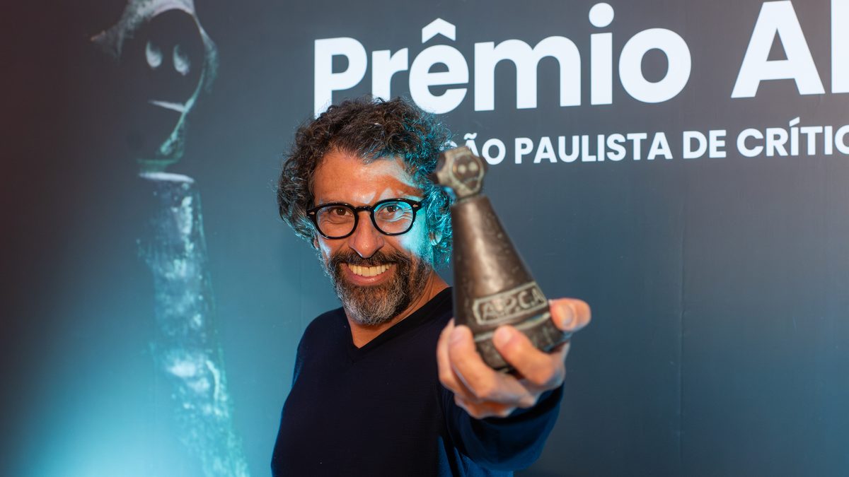 O ator Milhem Cortaz, o Juarez de Por Você, sorri e estende um troféu na cerimônia do Prêmio APCA, em São Paulo, em julho de 2024