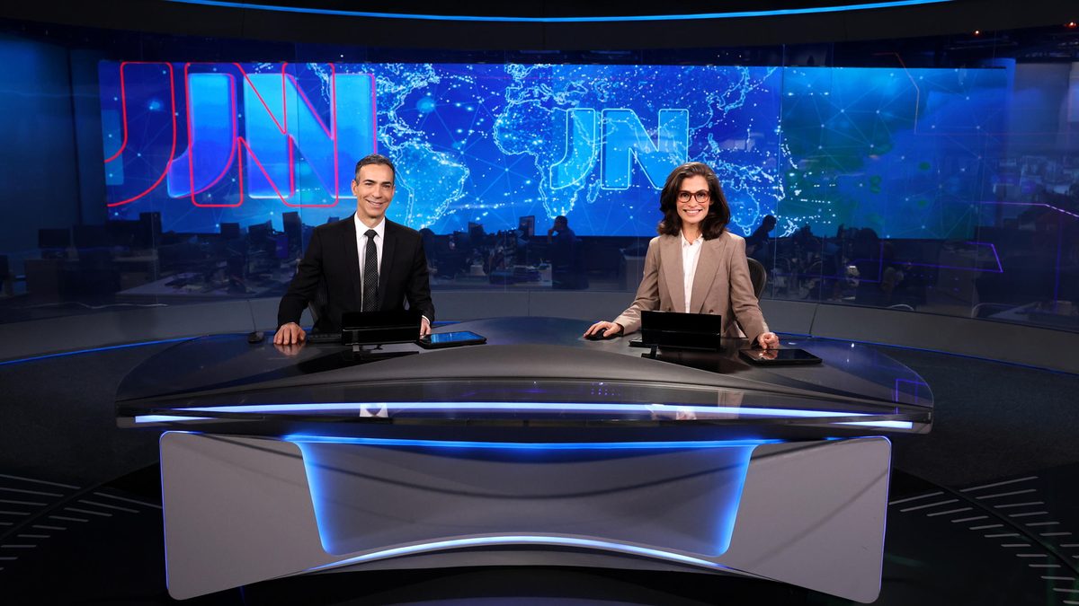 César Tralli e Renata Vasconcellos sorriem sentados à bancada do estúdio do Jornal Nacional, em foto de divulgação da Globo