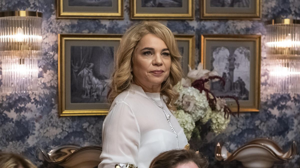 Mulher loira de blusa branca com lantejoulas, em pé entre convidados de um jantar, em cena de Quem Ama Cuida