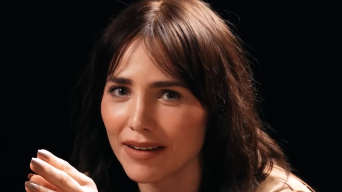 A atriz Letícia Colin, de cabelo castanho e blusa bege, gesticula com a mão diante de um fundo preto