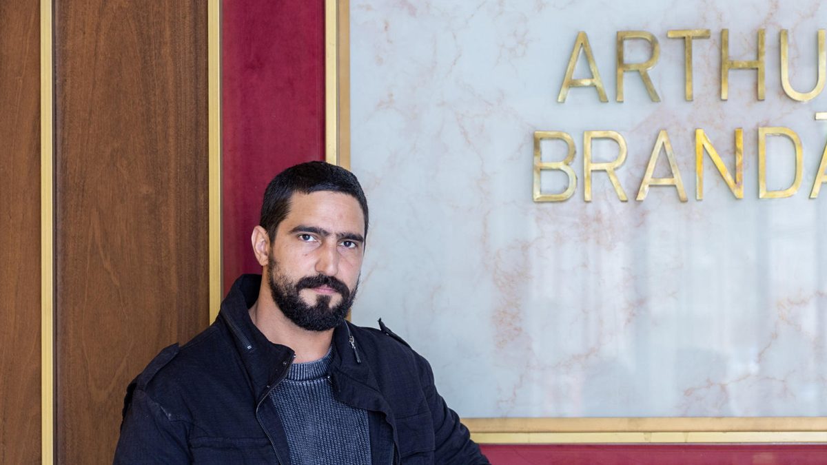 Homem de barba e jaqueta escura diante de um painel dourado com o nome Arthur Brandão, em foto de divulgação de Quem Ama Cuida