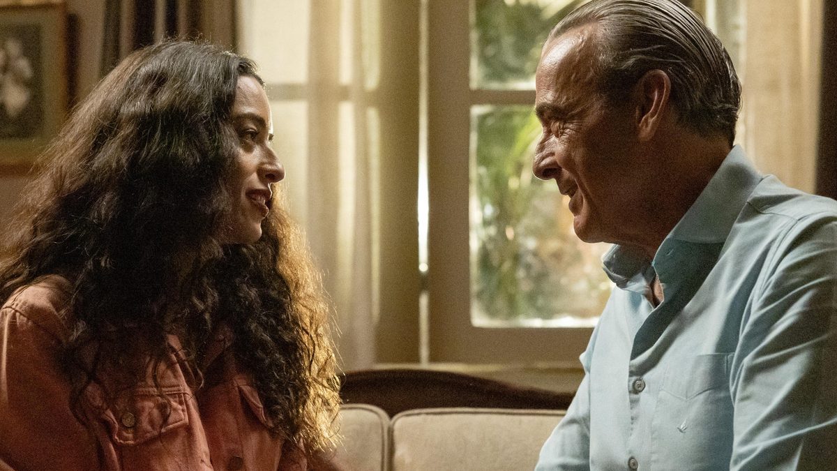 Lyris, de camisa salmão e cabelo cacheado, e Ulisses, de camisa azul-clara, conversam sorrindo num sofá em cena de Quem Ama Cuida
