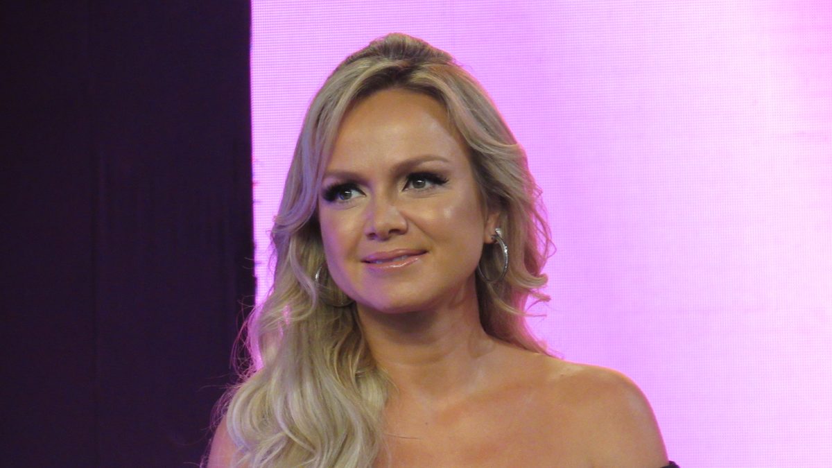 Eliana sorrindo, de cabelo loiro e longo e vestido preto, diante de um painel rosa, em foto de outubro de 2016