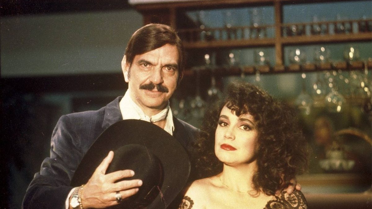 Homem de bigode segurando um chapéu preto e mulher de cabelo cacheado e vestido de renda posam juntos em foto de divulgação de Roque Santeiro