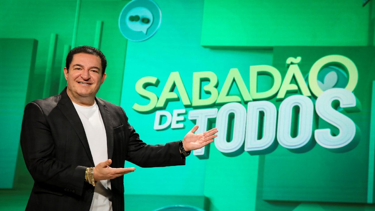 Renato Ambrósio, de blazer preto e camiseta branca, apresenta o logo do Sabadão de Todos no cenário verde do programa, no SBT