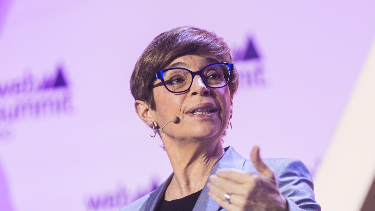 A jornalista Renata Lo Prete, de óculos e terno azul-acinzentado, gesticula enquanto fala sentada em um palco, em evento de 2024
