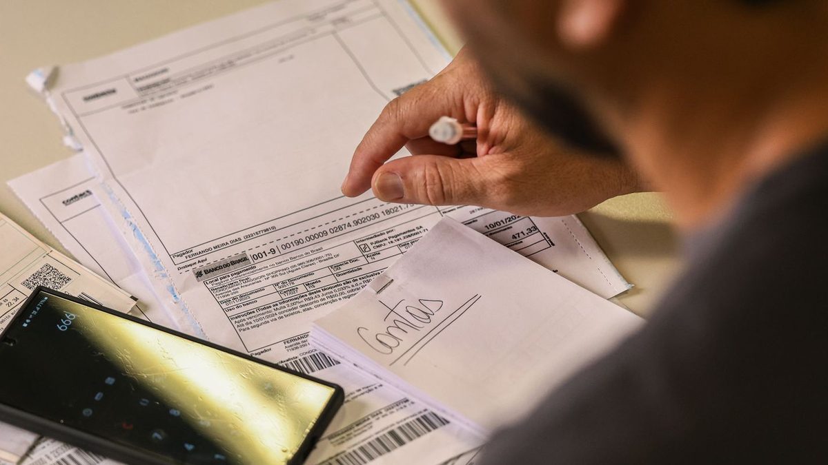 Mãos de uma pessoa conferindo boletos e contas de consumo sobre uma mesa, com um papel escrito Contas