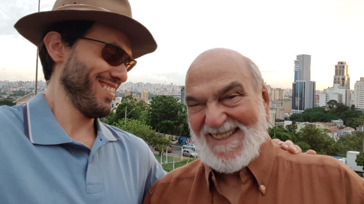 O ator Lima Duarte, de barba branca e camisa marrom, sorri ao lado de um homem de chapéu e óculos escuros, com a cidade ao fundo