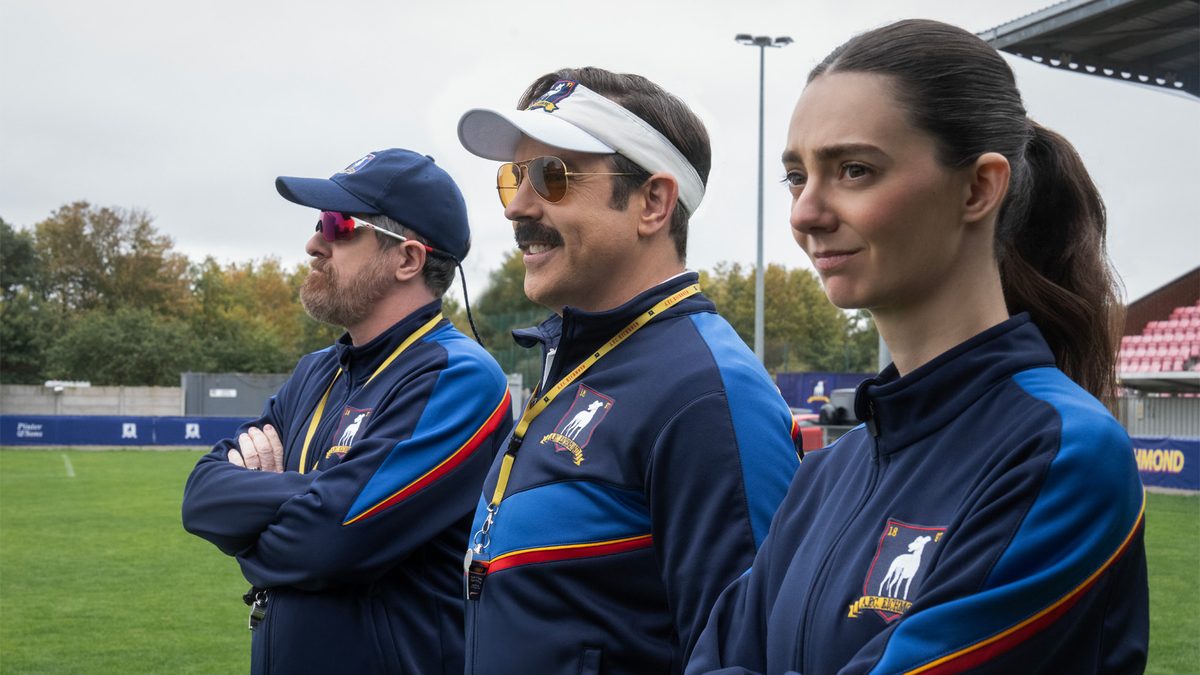 Brendan Hunt, Jason Sudeikis e Tanya Reynolds, de agasalho azul do AFC Richmond, ao lado de um campo, em foto de divulgação da 4ª temporada de Ted Lasso
