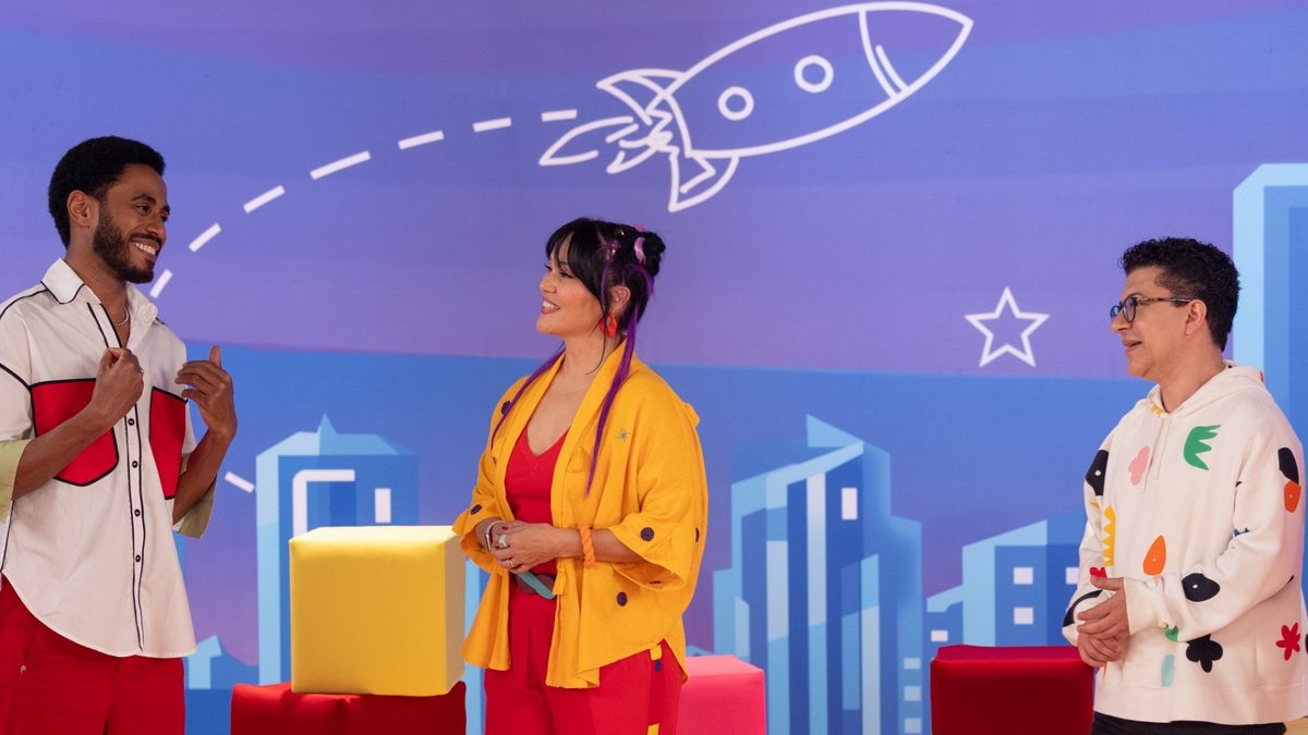 Apresentadores da edição especial da TV Globinho conversam em pé no cenário colorido do programa, com foguete e prédios desenhados ao fundo