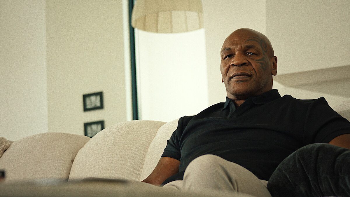 Mike Tyson, de camisa polo preta, sentado num sofá claro em imagem de divulgação da série documental TYSON, da Netflix