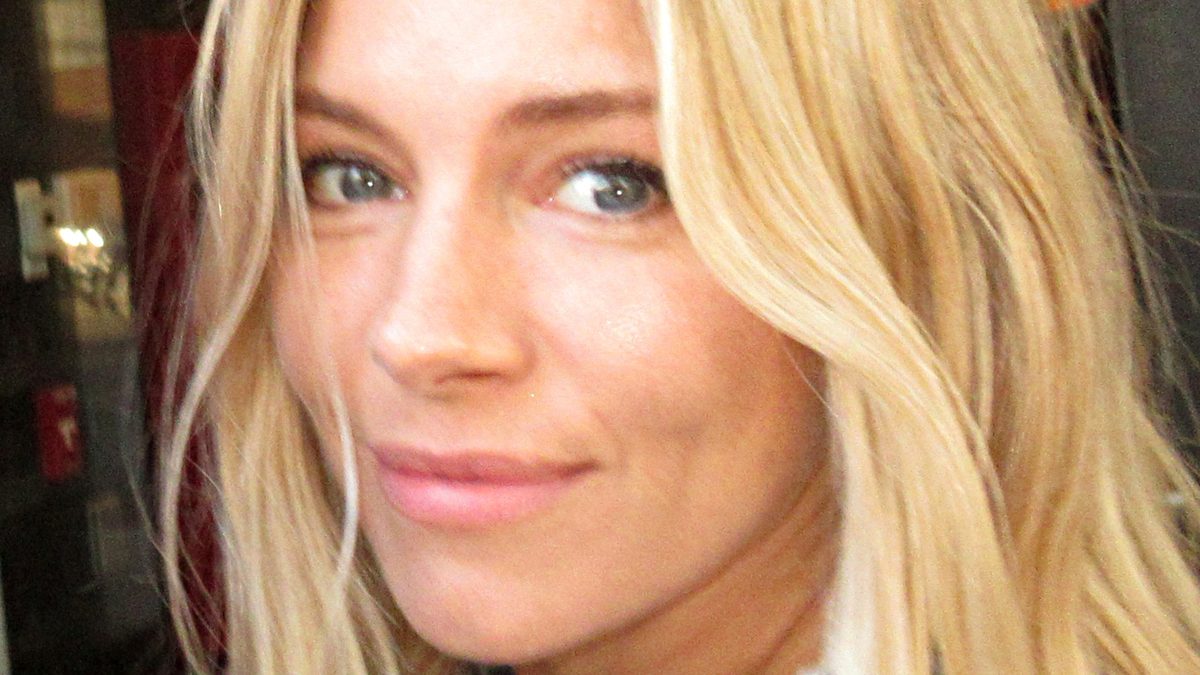 A atriz Sienna Miller, de cabelo loiro solto e blusa florida, sorri de lado em Nova York, em 2018